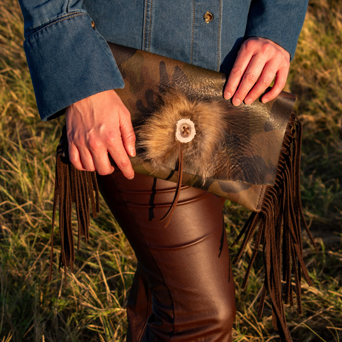 A woman stands in a grassy field at sunset wearing a blue denim shirt and brown leather pants, holding a leather clutch handbag with fringe, fur and an antler button..