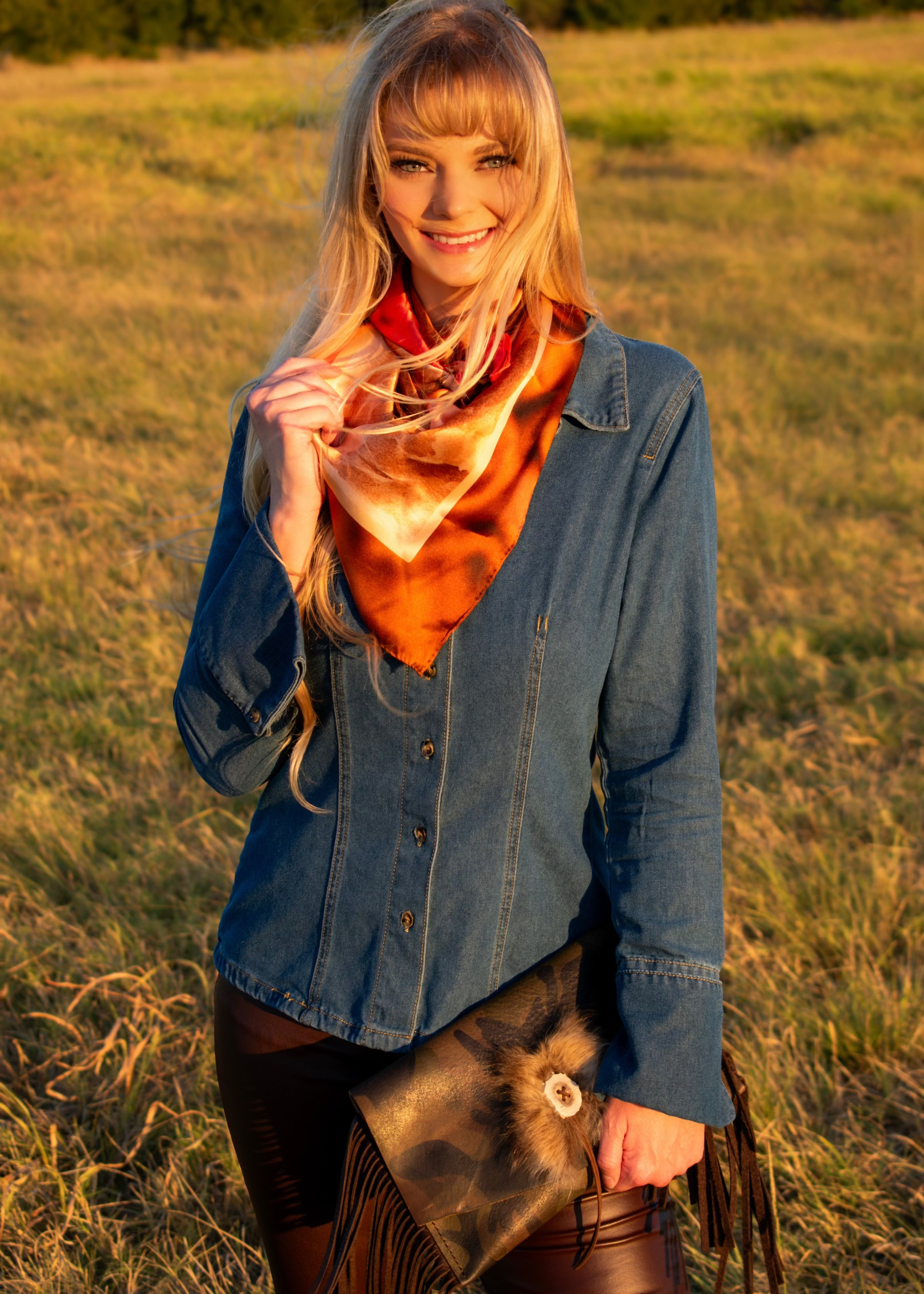 Woman in a denim shirt wearing the Absaroka silk scarf at golden hour, carrying a Bridger Leather camo clutch with fringe
