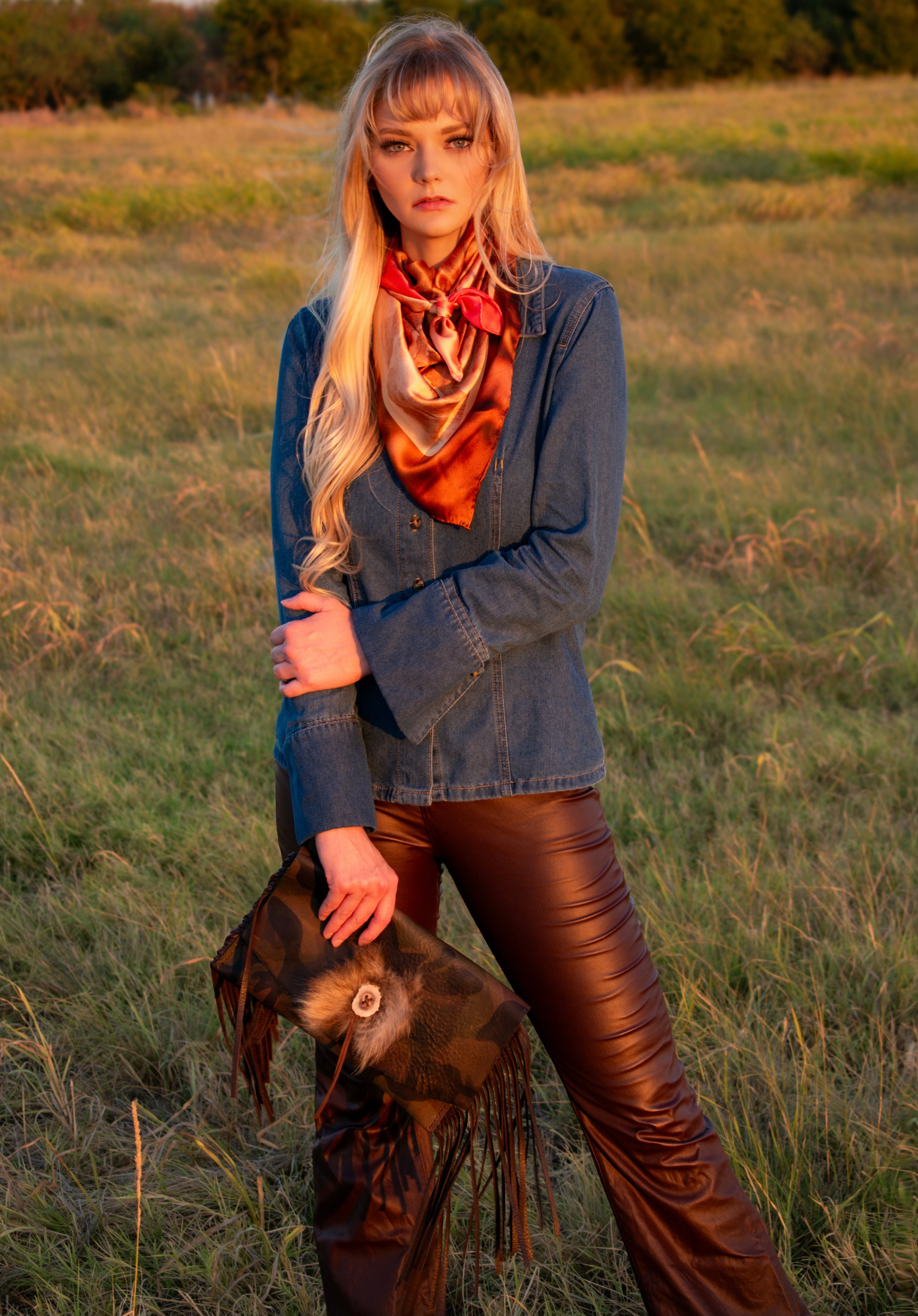 A woman stands in a grassy field at sunset wearing a blue denim jacket, a rust-orange patterned scarf, brown leather-look pants, and holding a clutch handbag.