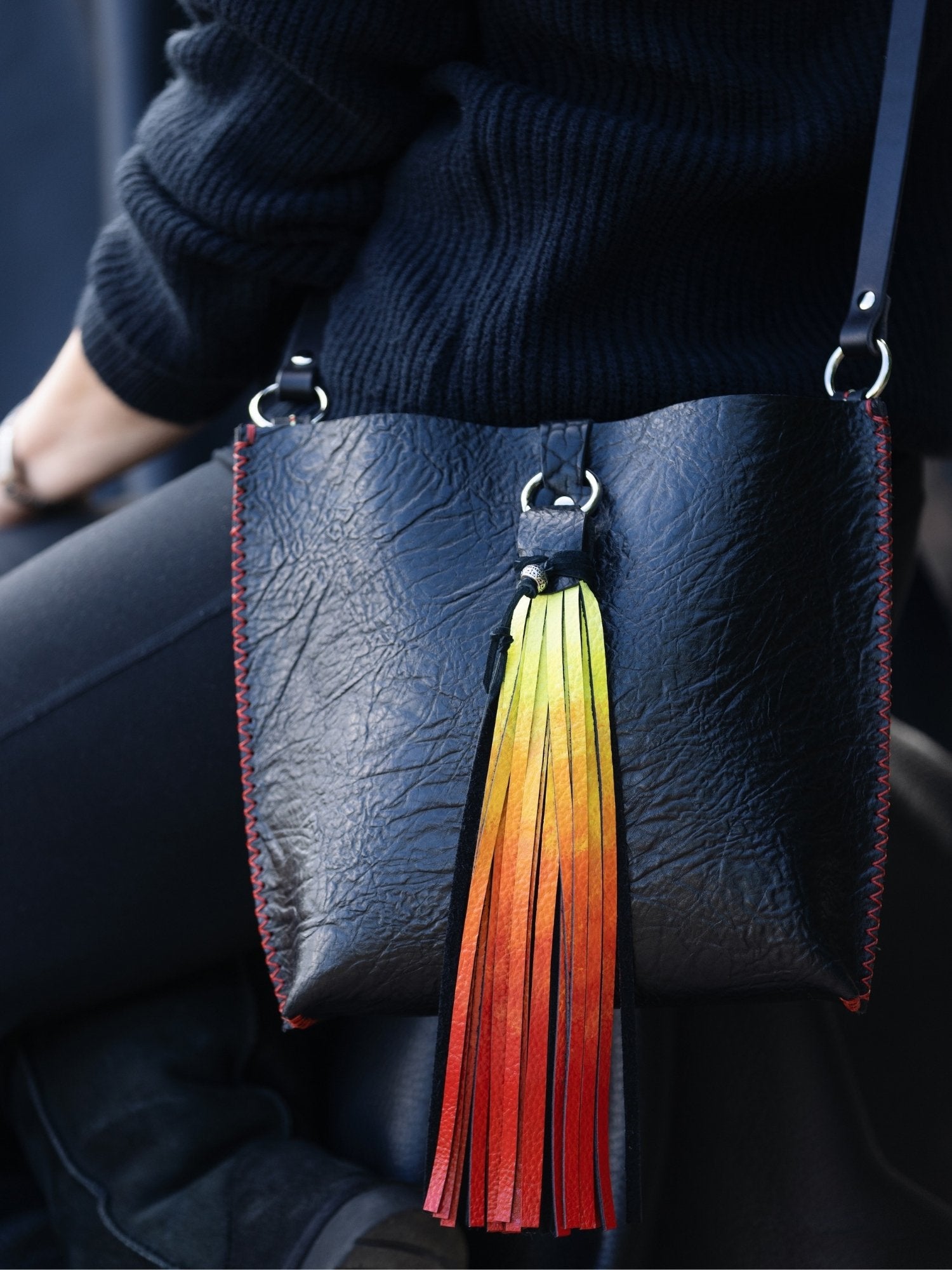 A close-up of a person wearing a black ribbed sweater and black pants, carrying a black leather shoulder bag with a long tassel charm that fades from yellow to orange to red.