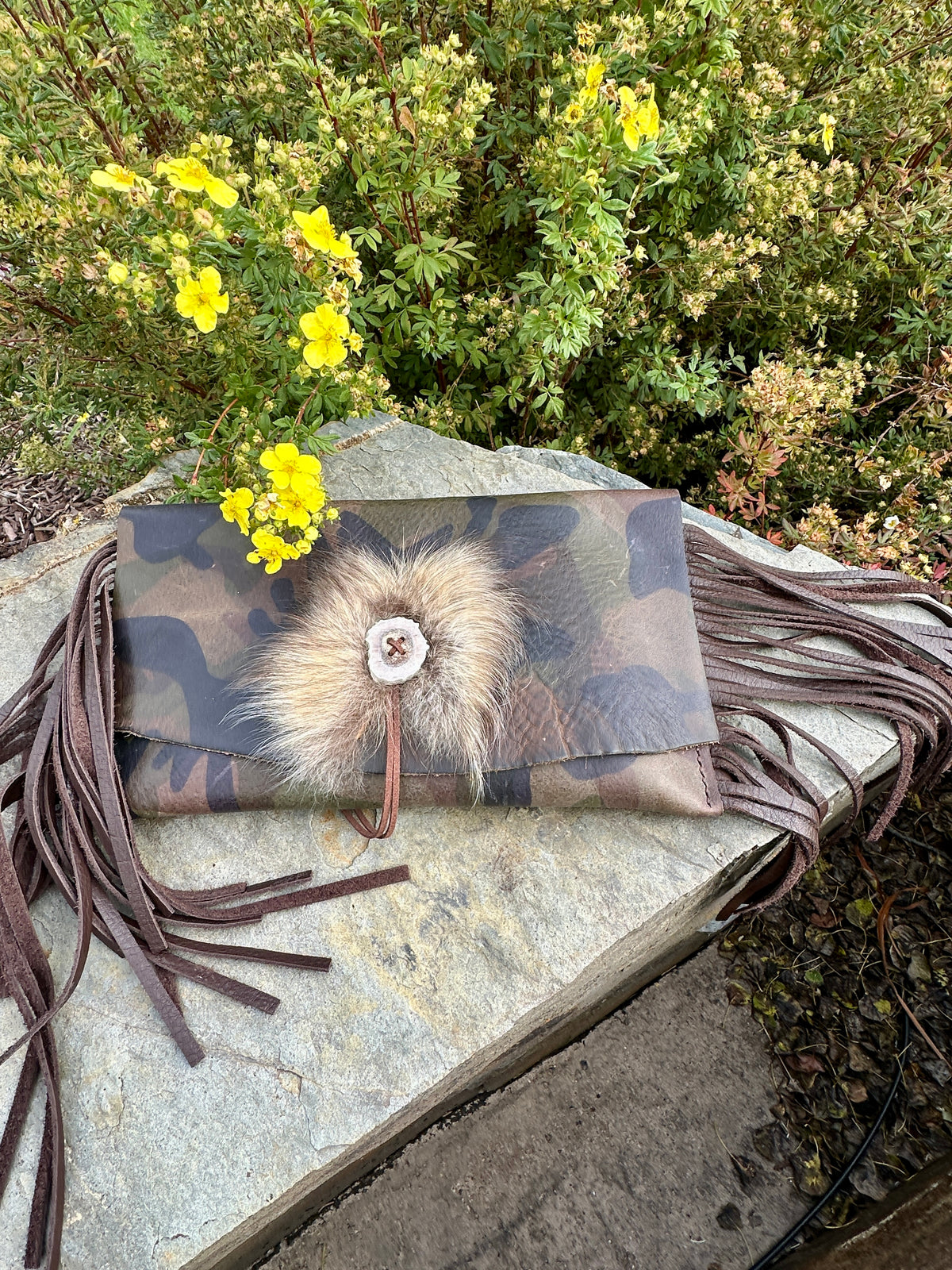 Close view of the Upland Camo Leather Clutch by Bridger Leather, showing the antler button and Finn raccoon fur accent on the camo-printed leather flap with leather fringe