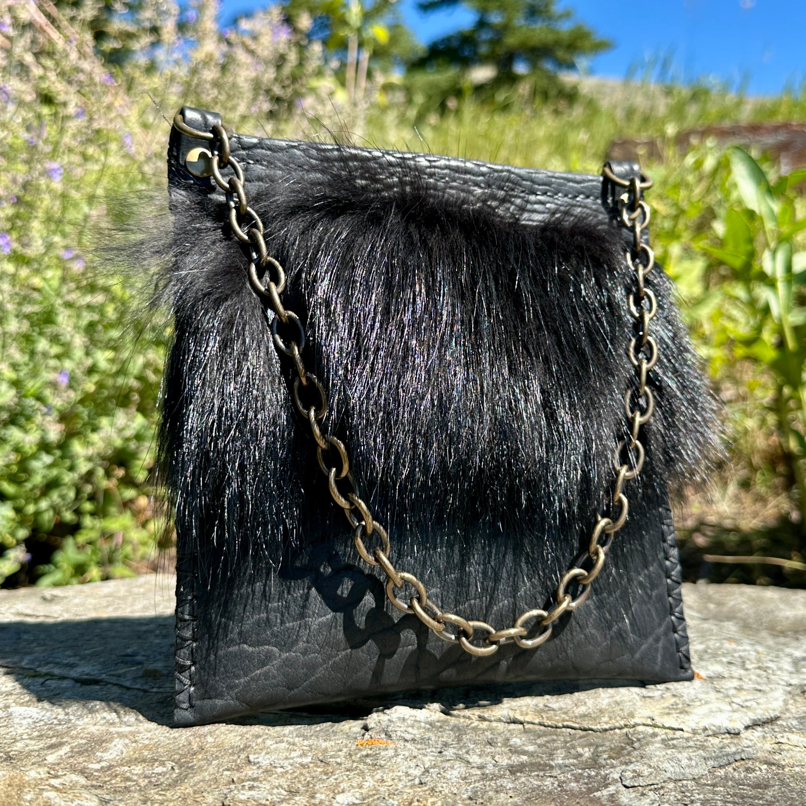The Ana Evening Bag in black bison leather with black coyote fur trim and an antique brass chain strap, resting on a stone outdoors