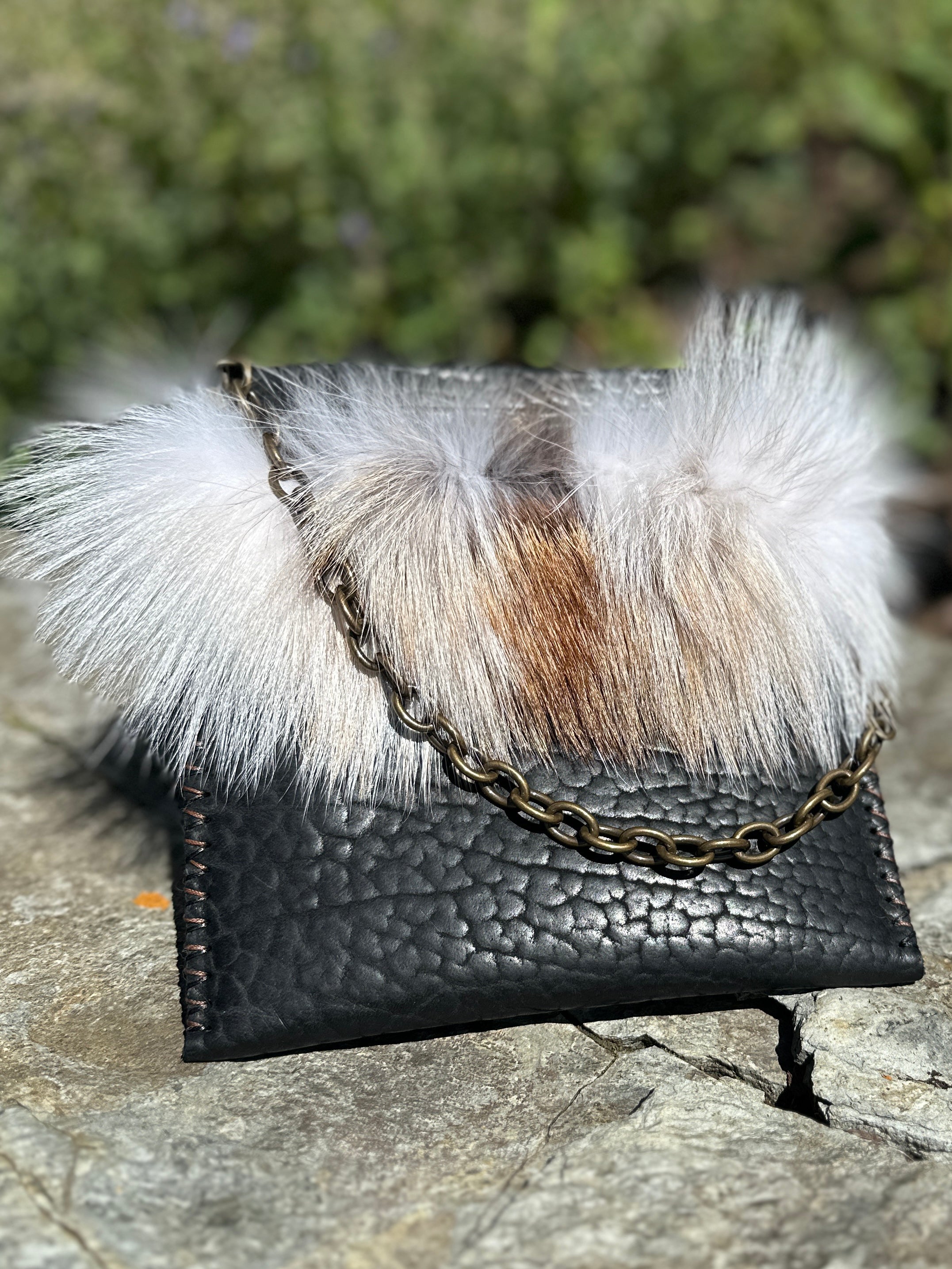 Front angle of the Ana Evening Bag with fox fur trim and antique brass chain resting on a stone outdoors