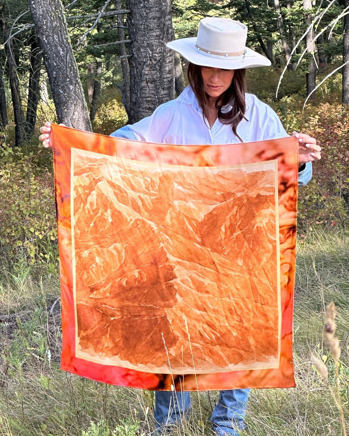Woman in a cowboy hat holding the Absaroka silk scarf open in the woods to show its full 35 inch print