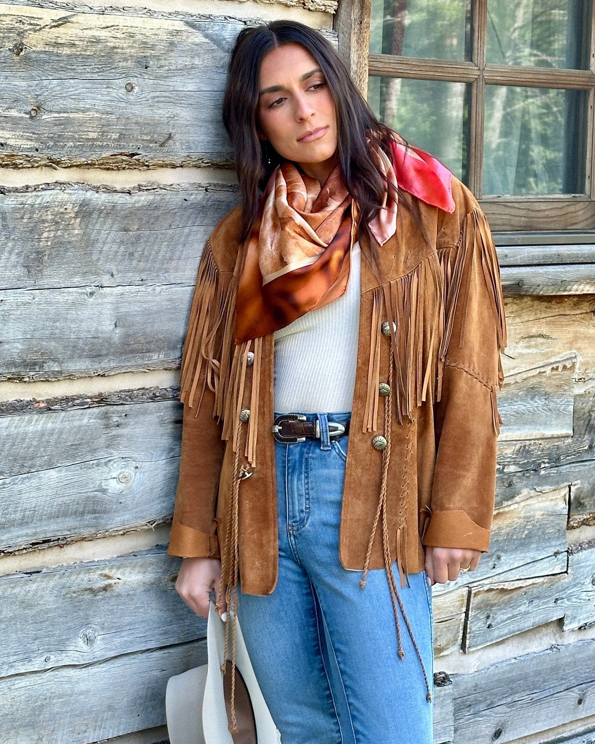 Woman in a fringed suede jacket and jeans wearing the Absaroka silk scarf, leaning against a log cabin wall