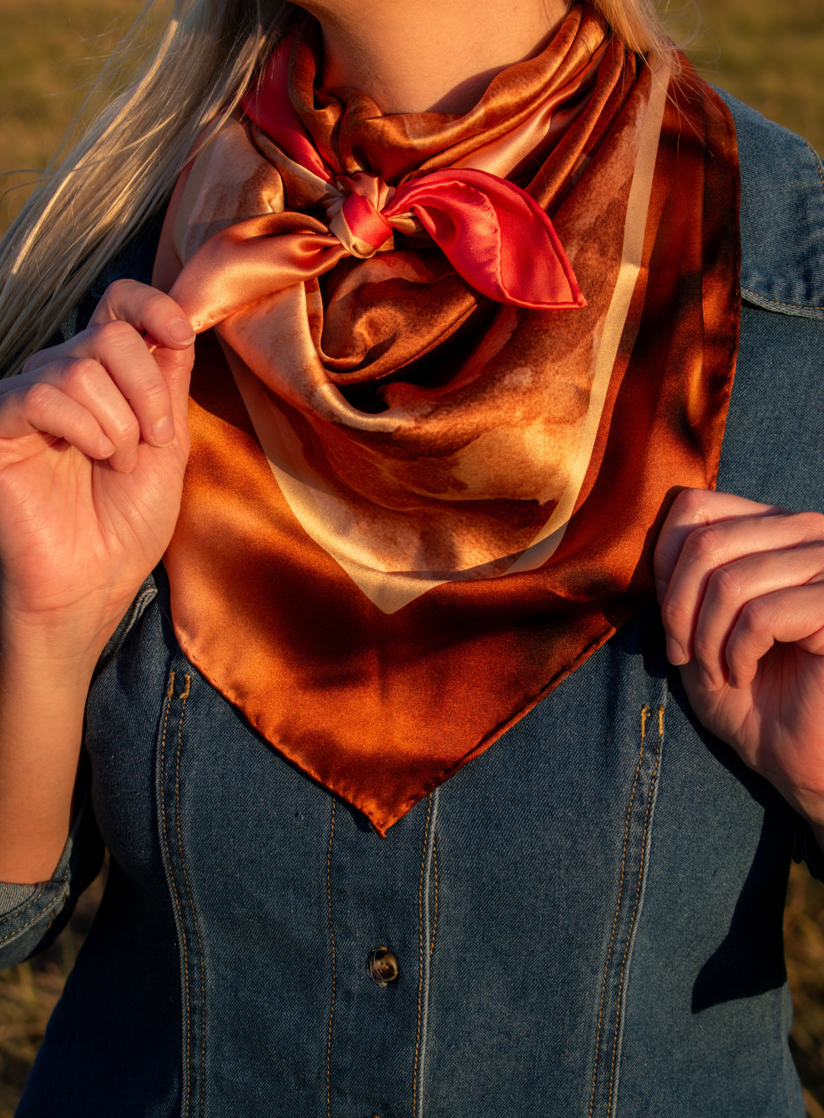 A close-up of a person wearing a satin scarf in warm rust orange, bronze, and red tones, tied in a bow at the neck over a blue denim top, with outdoor light and a blurred natural background.