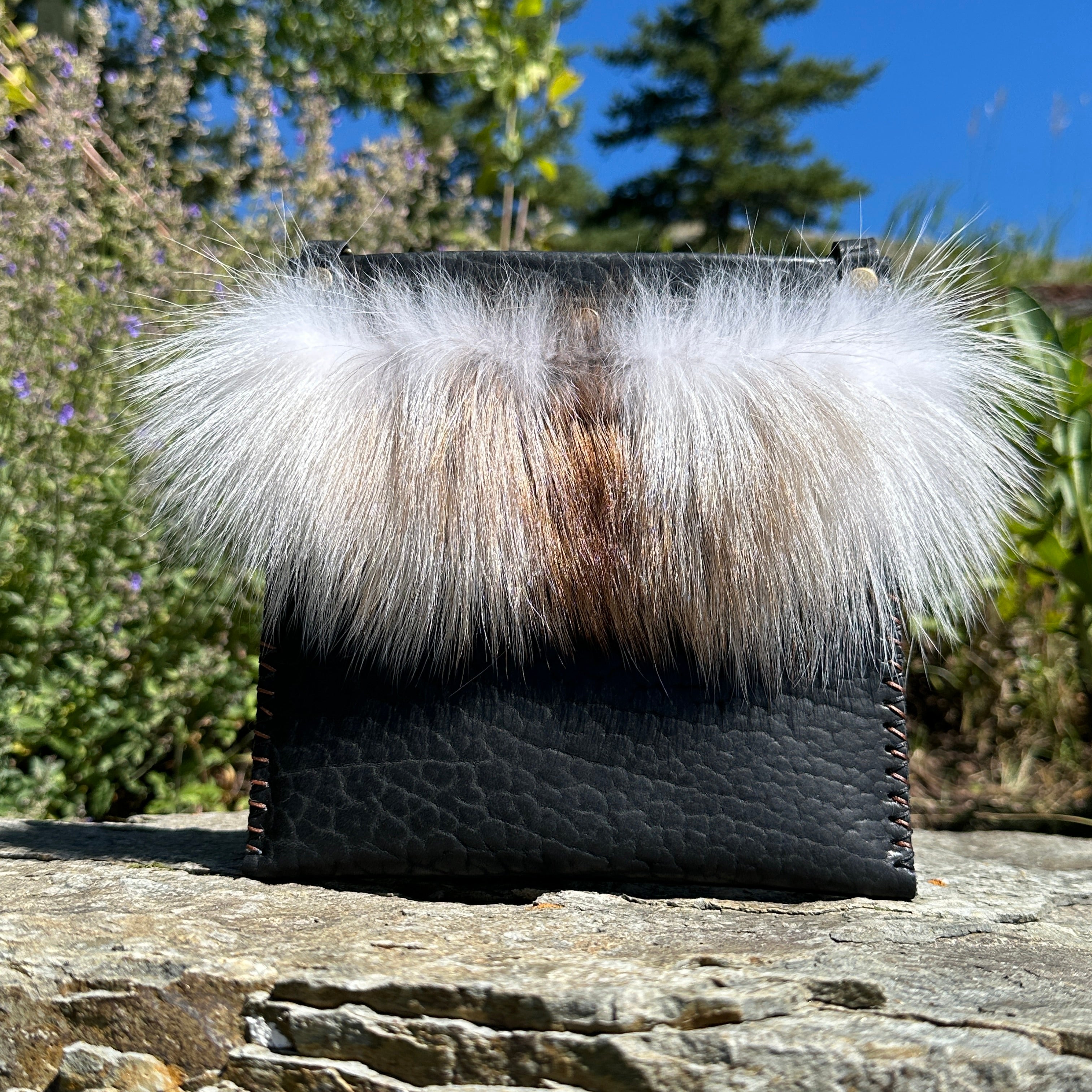 The Ana Evening Bag in black bison leather with natural fox fur trim and an antique brass chain, resting on a stone outdoors