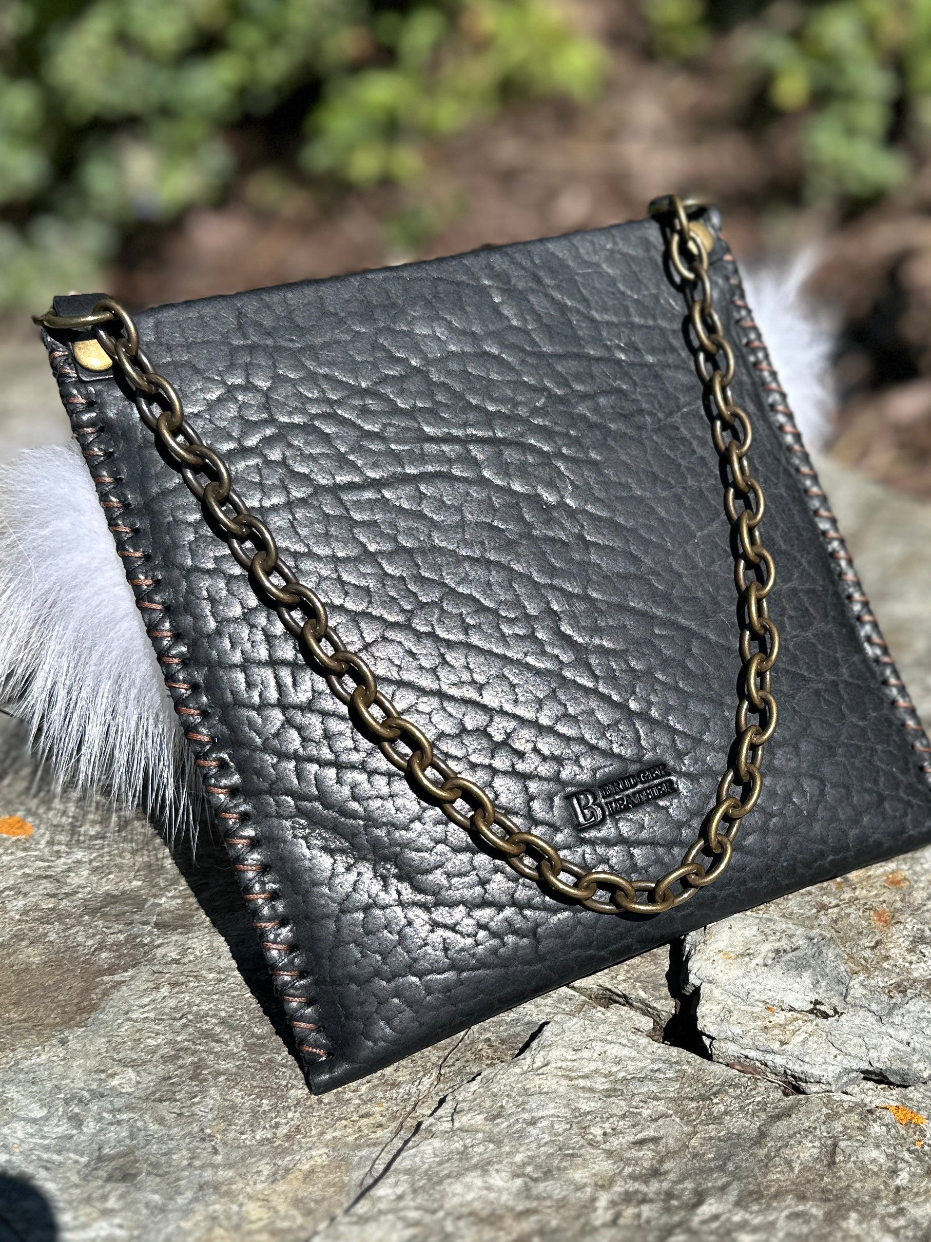 Back of the Ana Evening Bag showing the pebbled black bison leather, antique brass chain, and Bridger Leather logo stamp, with fox fur trim visible at the sides