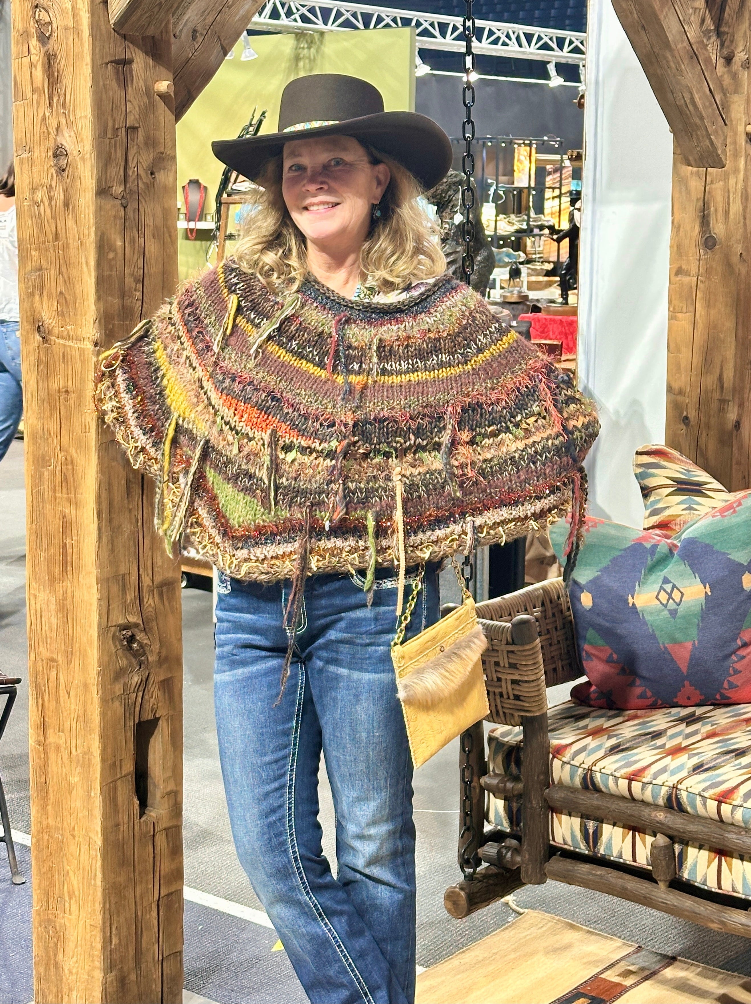 A woman wearing a brown and black striped knit poncho with fringe details, blue jeans with embroidered back pockets, and a black cowboy hat, standing in front of a large tree trunk.