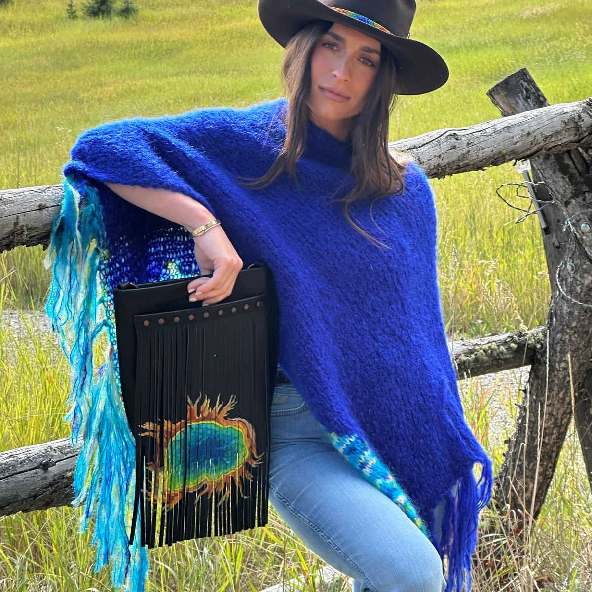 A woman outdoors in a grassy field leans against a wooden fence, wearing a bright royal blue fuzzy poncho with turquoise fringe, a dark brown wide-brim hat, and light blue jeans. She holds a black fringed handbag with a colorful painting of the grand prismatic spring design.