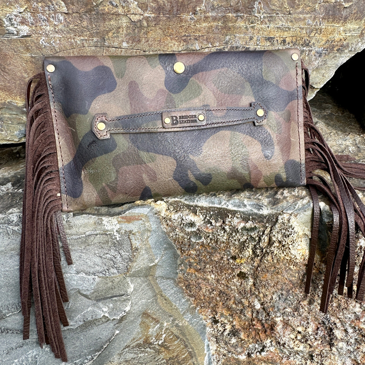 Flat front view of the Upland Camo Leather Clutch by Bridger Leather, showing the camo-printed leather, strap closure with Bridger Leather hardware, and long leather fringe on both sides