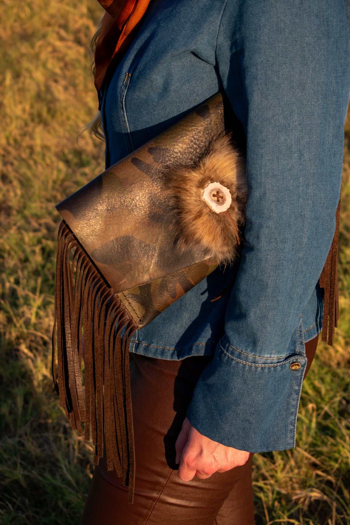 A close-up lifestyle shot of a person outdoors wearing a brown leather fringe clutch bag with a fur trim and a white antler button detail, paired with a blue denim jacket and brown leather pants.